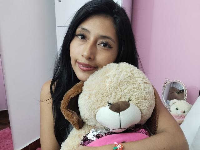 LucianaHernandez with long black hair, holding a fluffy beige teddy bear, smiling at the camera, wearing pink lipstick, and a black sleeveless top, in a pink room.