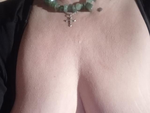 MioritaStar's bare chest with visible cleavage, wearing a green bead necklace with a silver cross, showing light skin and small moles. Boobs, necklace, and beads are visible.