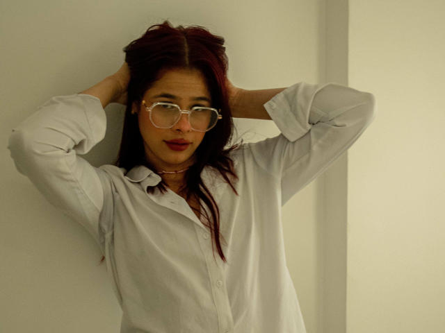 EuphoriaRush, com as mãos no cabelo ruivo escuro, vestindo uma camisa branca folgada e óculos transparentes, em pé contra uma parede branca, com uma expressão ligeiramente irritada. Seios não visíveis..