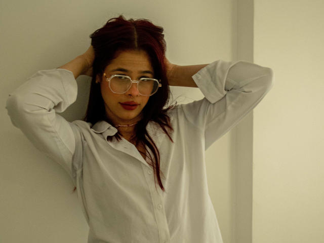 EuphoriaRush mit roten Haaren, Brille und weißem Hemd, Hände im Haar, leicht lächelnd, kleine Brüste, süß, lässig, stehend vor einer weißen Wand, entspannt, selbstbewusst..
