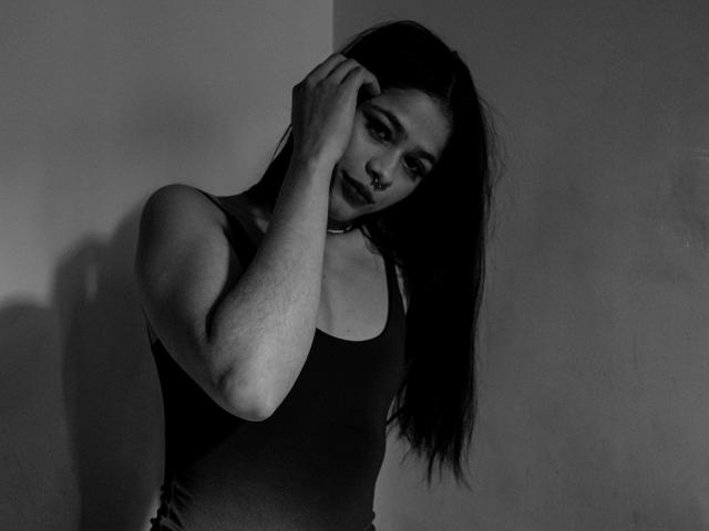 Foto in bianco e nero di EuphoriaRush con lunghi capelli scuri, che si tocca la testa, indossa una canotta aderente, mostra un braccio muscoloso e un piercing al naso, guarda in basso con un'espressione seria..