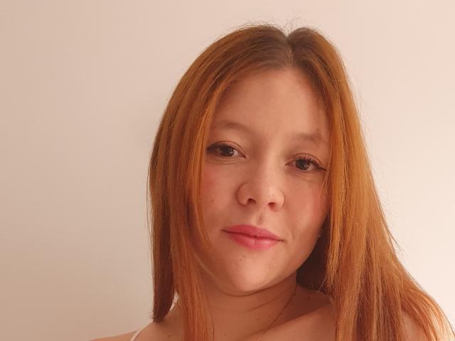 EuphoriaRush a les cheveux bruns clairs et lisses, une peau claire et des lèvres roses, avec un léger sourire. Ses yeux bruns sont légèrement en amande, et elle porte un haut blanc..