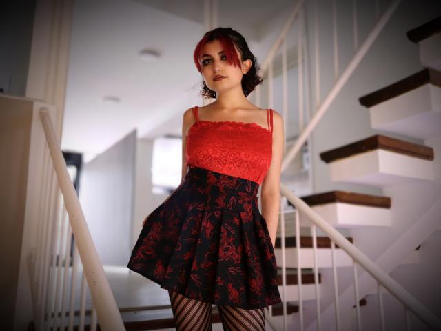 AngelBlakee with red lace top, black floral skirt, fishnet stockings, standing on staircase, short dark curly hair with red highlights, confident pose, small tits, cute face, white background.