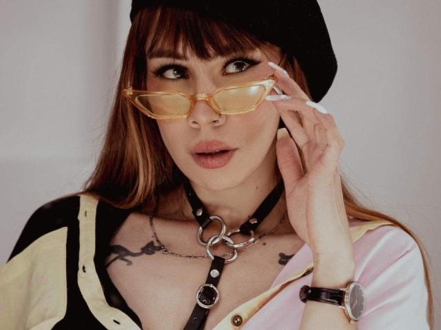 Ariianna, belle, avec des lunettes, un chapeau noir et un collier en cuir, tenant son téléphone, montrant des tatouages sur sa poitrine, et portant un haut rose et jaune, a l'air séduisante..