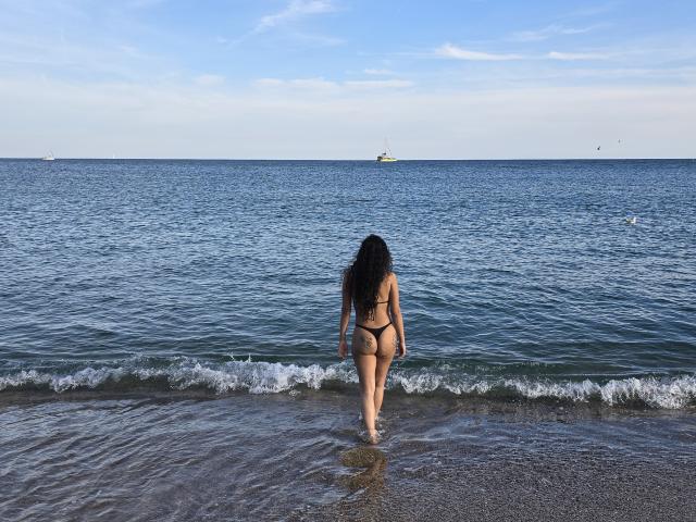 AlejandraMontana with long black hair, wearing a black thong bikini, stands in shallow water, her ass and back visible, facing the blue ocean under a clear sky.