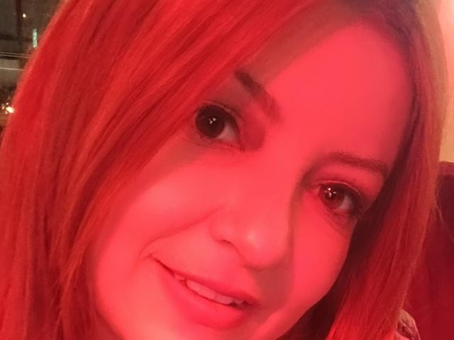 NatalyHami's face with red lighting, smiling with straight red hair, brown eyes, and visible eyebrows. Her nose and lips are slightly pink due to the red light.
