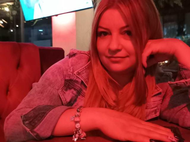 NatalyHami with red-pink lit face, long blonde hair, denim jacket, resting chin on hand, wearing a bracelet, smiling slightly, in a red tufted booth, looking cute.