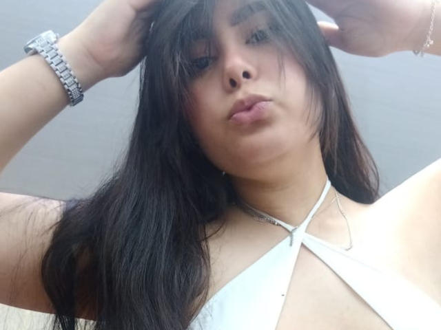 BrianaRogers with long dark hair, pouty lips, wearing a white halter top, showing small tits, silver bracelet, and a delicate necklace, looking down at the camera.