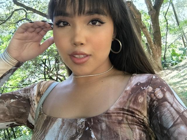 Heyly, with long black hair and bangs, has her hand near her head, wearing a brown tie-dye top and hoop earrings.