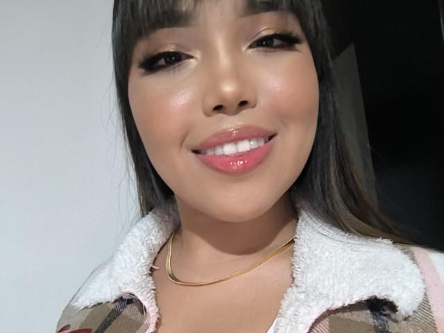 Heyly with glossy pink lips smiling, wearing a white-collared shirt, black hair with bangs, and a gold necklace.