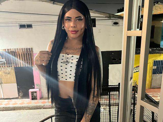 MarcelineDarketa has long black hair, tattoos on arms, a white crop top with black dots, and black pants, standing in a dimly lit room, with a confident smile.