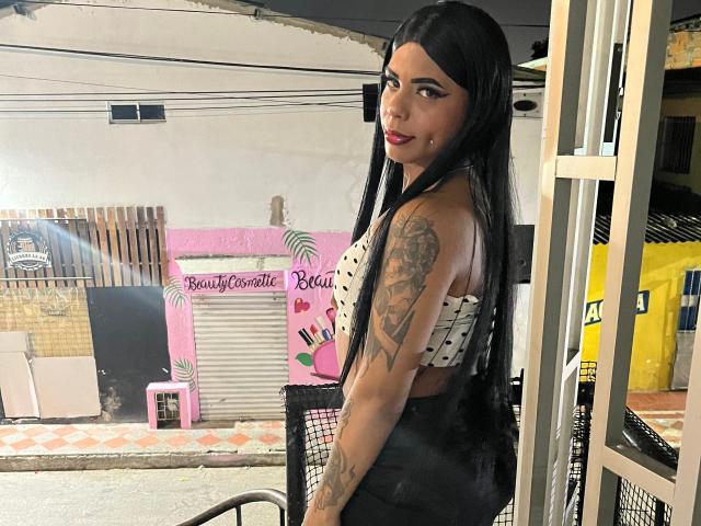 MarcelineDarketa with long black hair, white polka-dot crop top, black high-waisted skirt, tattooed arm, smiling at camera, standing in front of metal chair, in dimly lit room.