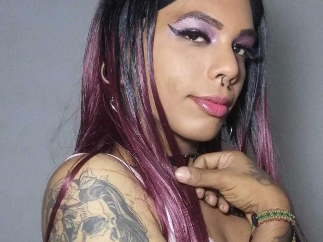 MarcelineDarketa has purple and black dyed hair, purple eyeshadow, nose ring, tattoo on right shoulder, pink lips, and a green bracelet, with a hand near her chin.
