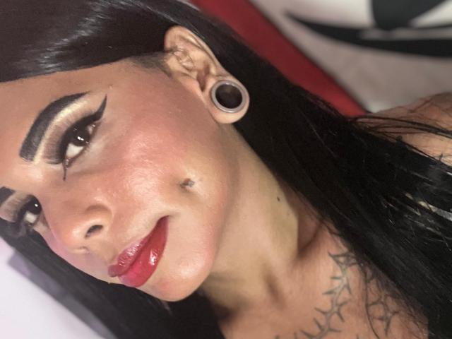 MarcelineDarketa with dark hair, bold eyeliner, red lips, ear gauges, small mole on her cheek, tattoo on her chest, and a slight smile, showing her glossy red lipstick.