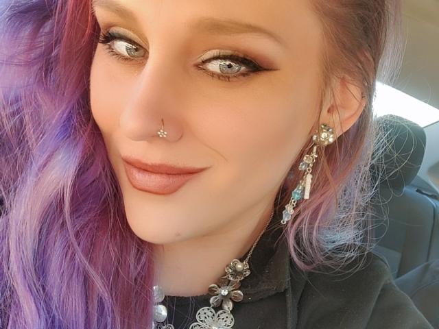 VikingBabe has purple and pink hair, a nose ring, and a septum piercing, with smoky eye makeup, pink lipstick, and floral earrings, sitting in a car.