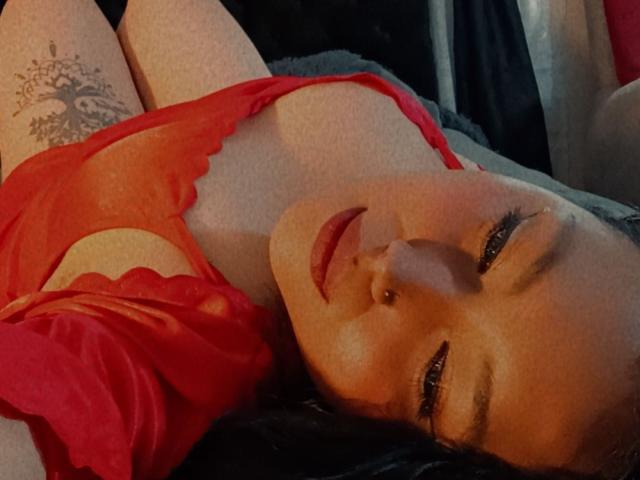KerstinJanell is lying down in red lingerie, showing her tits through the lace. She has a nose piercing and a tree tattoo on her right thigh.