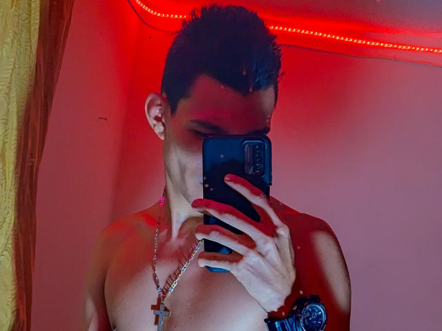 TheNewMan senza camicia, con capelli corti e scuri, smalto rosso sulle unghie, una croce al collo e un elastico al polso, che si fa un selfie allo specchio con una striscia di luci LED rosse sullo sfondo..