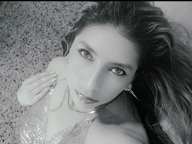 MichelleDumont with shiny sequin top, nose ring, hoop earrings, and glossy lips, looking up with hand near face, lying on speckled surface, black-and-white photo.