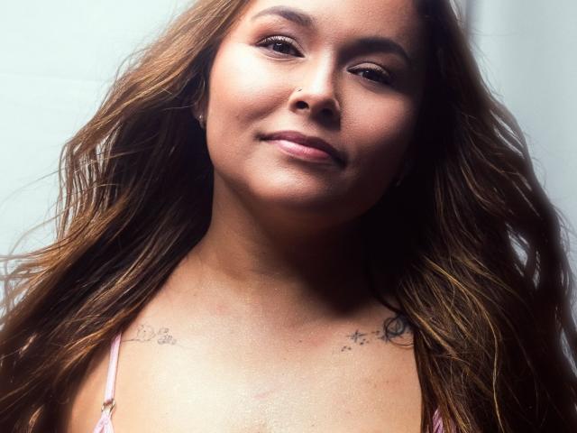 PinkyHotLover a la peau brun clair, des cheveux bruns ondulés, un anneau au nez et des tatouages sur le haut de sa poitrine. Elle sourit, a de petites seins et porte un haut rose..