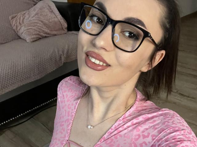 MonaLiss with black glasses, pink leopard-print top, and pink lipstick, smiling at camera. She's wearing a pearl necklace, and has white circles on her glasses.