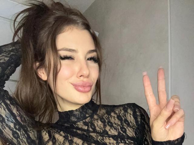 SensualMila with brown hair, black lace top, winking, and pouting, making a peace sign with her right hand, showing off long, manicured nails.