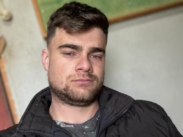 HotCoupleSapeca, a bearded dude with short brown hair, wearing a black jacket over a gray shirt, looking slightly annoyed with a subtle smirk, and a slightly flushed face.