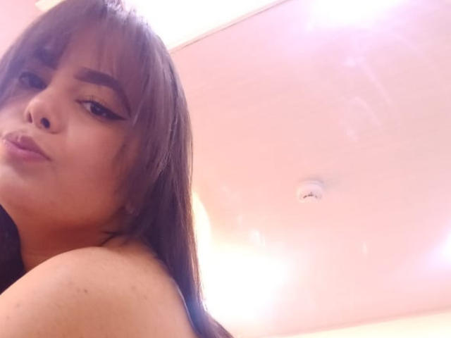 BrianaRogers with brown hair and bangs, pouty lips, and winged eyeliner, topless, showing her bare shoulders and upper chest, looking at the camera.