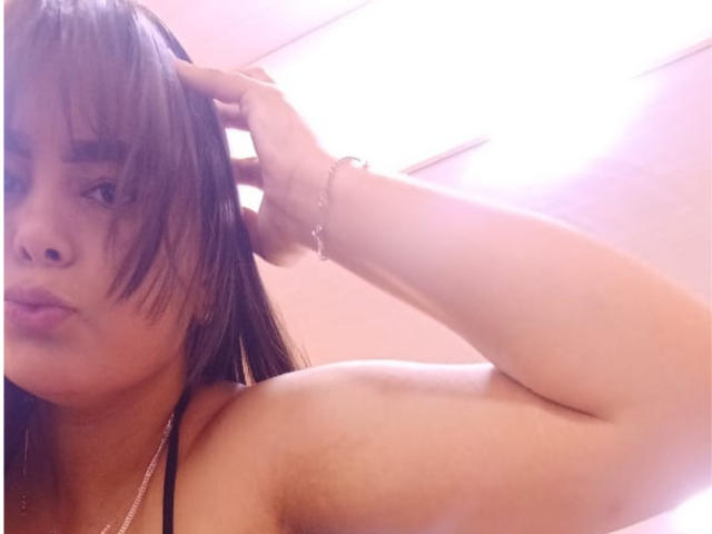 BrianaRogers with shoulder exposed, brown hair in face, hand in hair, pinkish light, bracelet on wrist, pouty lips, black strap on shoulder, soft light, slightly blurry.