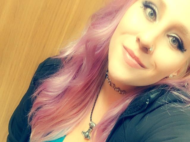 VikingBabe has pink-purple hair, blue eyes, black nose ring, black eyeliner, black choker, black jacket, and a necklace with a butterfly pendant. She's smiling at the camera.