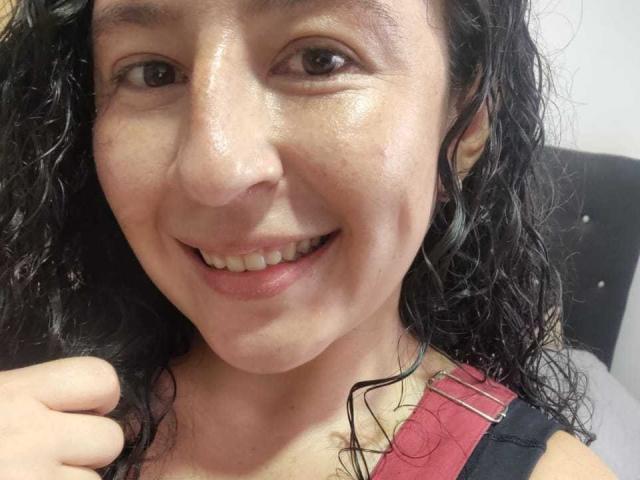 SweetyHot69 has a shiny, slightly sweaty face, dark curly hair, and a cute smile showing small teeth. She's wearing a red strap over her black top.