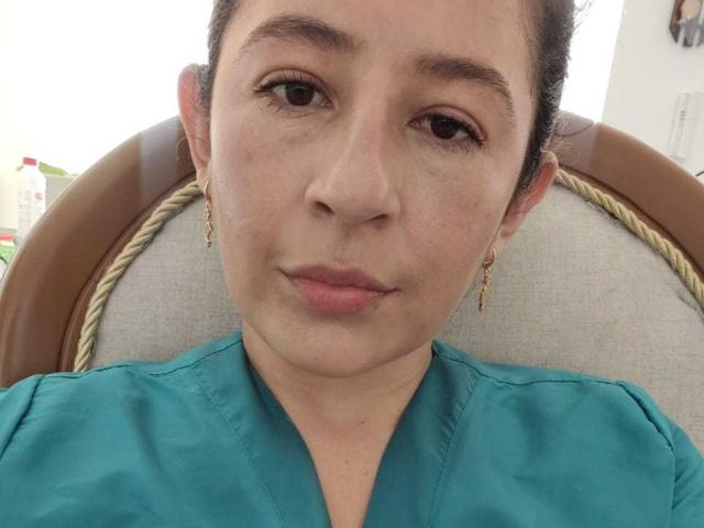 SweetyHot69 with brown eyes, medium skin, and dark hair in a bun, wearing teal shirt, gold earrings, slightly pouty lips, sitting on a brown chair with rope details.