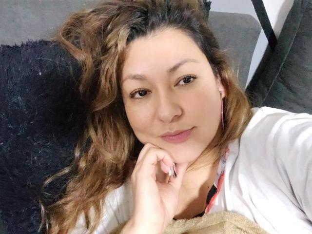 SakotoDika, con cabello castaño ondulado, piel clara, ojos marrones y aretes rosas, apoyando su barbilla en la mano, vistiendo una camisa blanca y sonriendo ligeramente..
