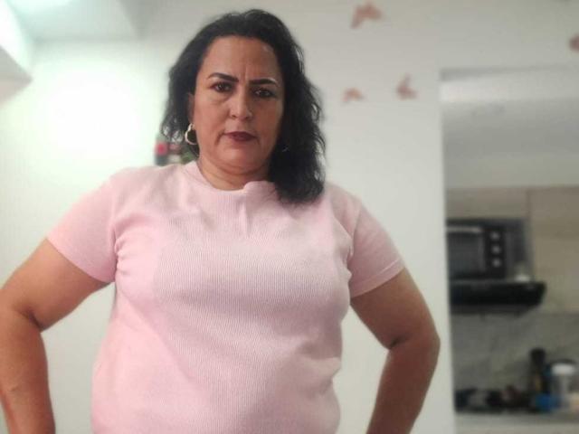 SaraHotLover, a chubby woman with curly black hair, wearing a pink ribbed t-shirt, standing with hands on hips, looking slightly annoyed, showing her tits.