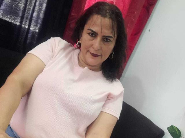 SaraHotLover with dark hair, pink shirt, small tits, hoop earrings, and dark lipstick, sitting on black couch, looking slightly annoyed. Red and black curtain in background.