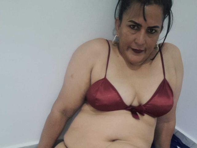 SaraHotLover, a chubby woman with dark hair in a red bra, sitting, showing her tits, a slight belly, and dark lipstick, looking at the camera.