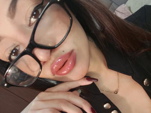 AnnaKhalo with glossy pink lips, black-framed glasses, and dark red nails, wearing a black buttoned shirt and gold necklace, touching her chin.