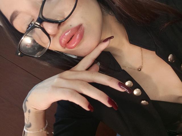 AnnaKhalo with glasses, puckered pink lips, finger touching them, dark red nails, black buttoned shirt, gold necklace, tattoo on hand, brown hair, wood background.