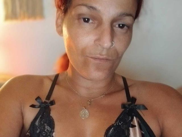 AigleHot with red hair, nose ring, and subtle smile, wearing black lace lingerie with ribbon accents, showing her tits and a necklace with a leaf pendant.