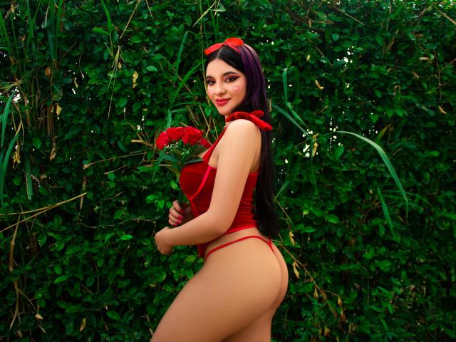 LylaHeather with long dark hair, red eyes, wearing red velvet top, thong, holding roses, standing sideways, showing ass, red sunglasses on head, smiling, green leafy background.