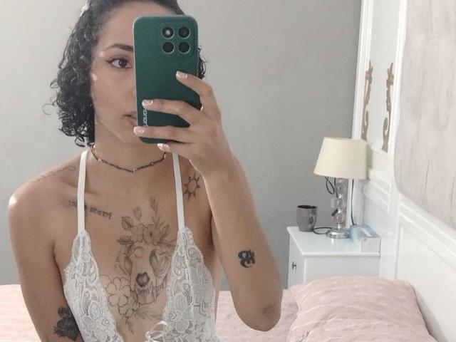 SilvanaXOwen with curly black hair, wearing a white lace halter top, showing tattoos on her chest and arm, holding a green phone, taking a mirror selfie.