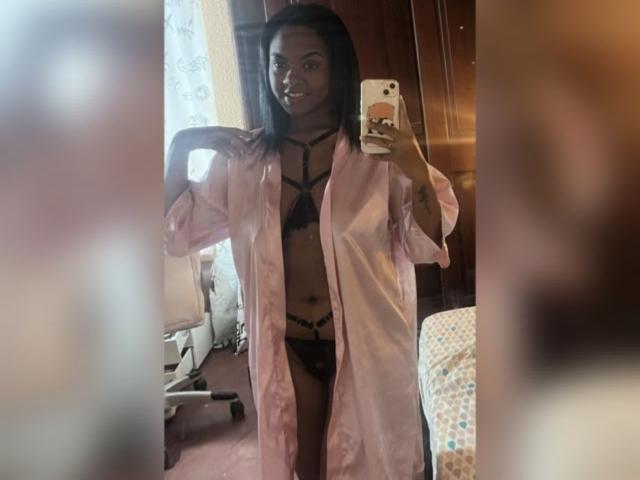 TiffanySammy with dark skin, black bra and panties, pink open shirt, holding phone in mirror, showing small tits and ass. She's smiling, with long black hair, in bedroom.