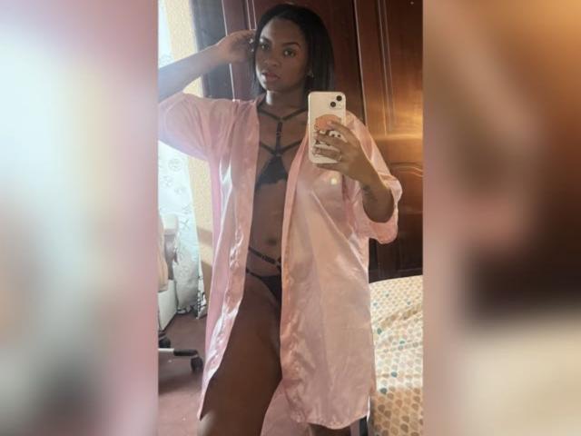 TiffanySammy in pink shirt, black lingerie, strappy bra, visible pussy, mirror selfie, holding phone, wooden door, bed with patterned sheets, brown carpet, reflective mirror.