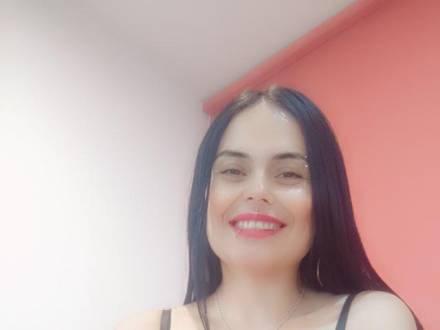 CharlotteCrofort has shiny black hair, a cute smile with red lipstick, and is wearing a black strap top. Her hoop earrings and glistening face are visible.