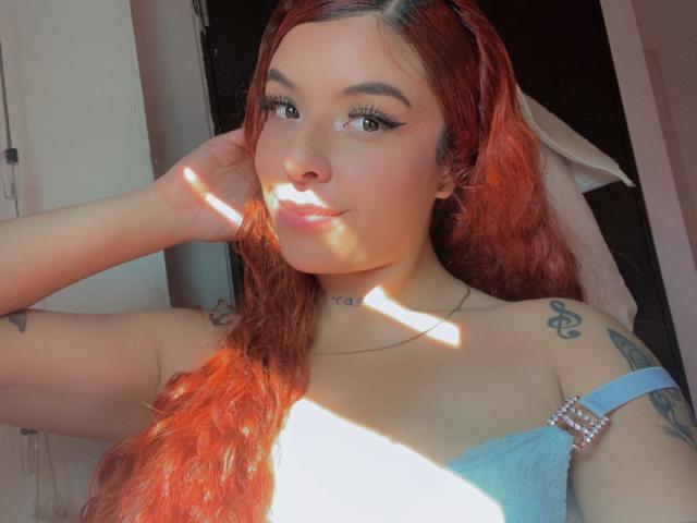 TammyyRoss, con capelli rossi, pelle chiara, tatuaggi sulle spalle, indossa un vestito blu con spalline, luce solare sul viso, una mano tra i capelli, un sorriso leggero, eyeliner e una collana..