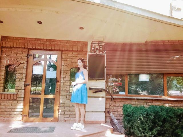 MirandaHott69, pregnant with a small baby bump, wears a blue dress and white sneakers, standing in front of a brick building with a wooden door and grill.