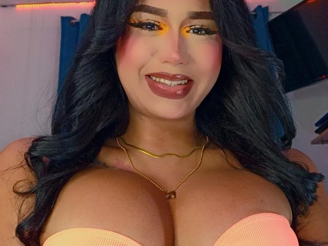 AnielFetish with big, orange-tinted eyelids, wavy black hair, and full pink lips, smiling at the camera, wearing a gold necklace, and a strapless orange top showing her tits.