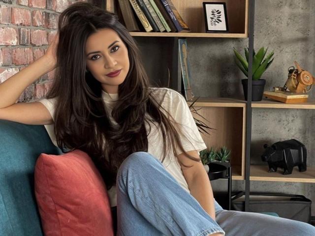 Loren Holmes, com cabelos longos e escuros, sentada em um sofá azul-turquesa, vestindo uma camiseta branca e jeans azuis, com a mão no cabelo, um travesseiro vermelho, uma estante com plantas e brinquedos ao fundo..