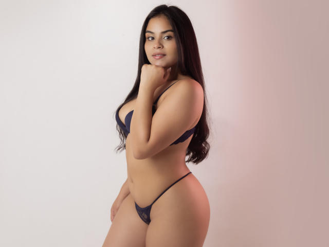 DanaCami with long dark hair, wearing a blue lace bra and thong, has a curvy figure, small boobs, and a slight smile, standing sideways with a hand on her neck.