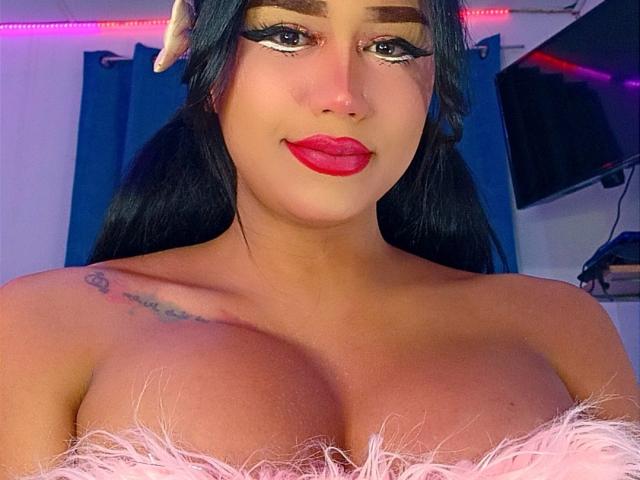 AnielFetish with big tits, red lipstick, black hair, white hair clip, tattoo on left shoulder, white furry strap, blue curtains, and a TV in the background.