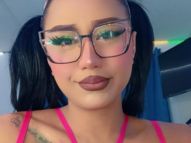 AnielFetish has dark puffy twin pigtails, green eyeshadow, thick black-framed glasses, pink top, brown lipstick, tattoo on left shoulder, and a small nose piercing.